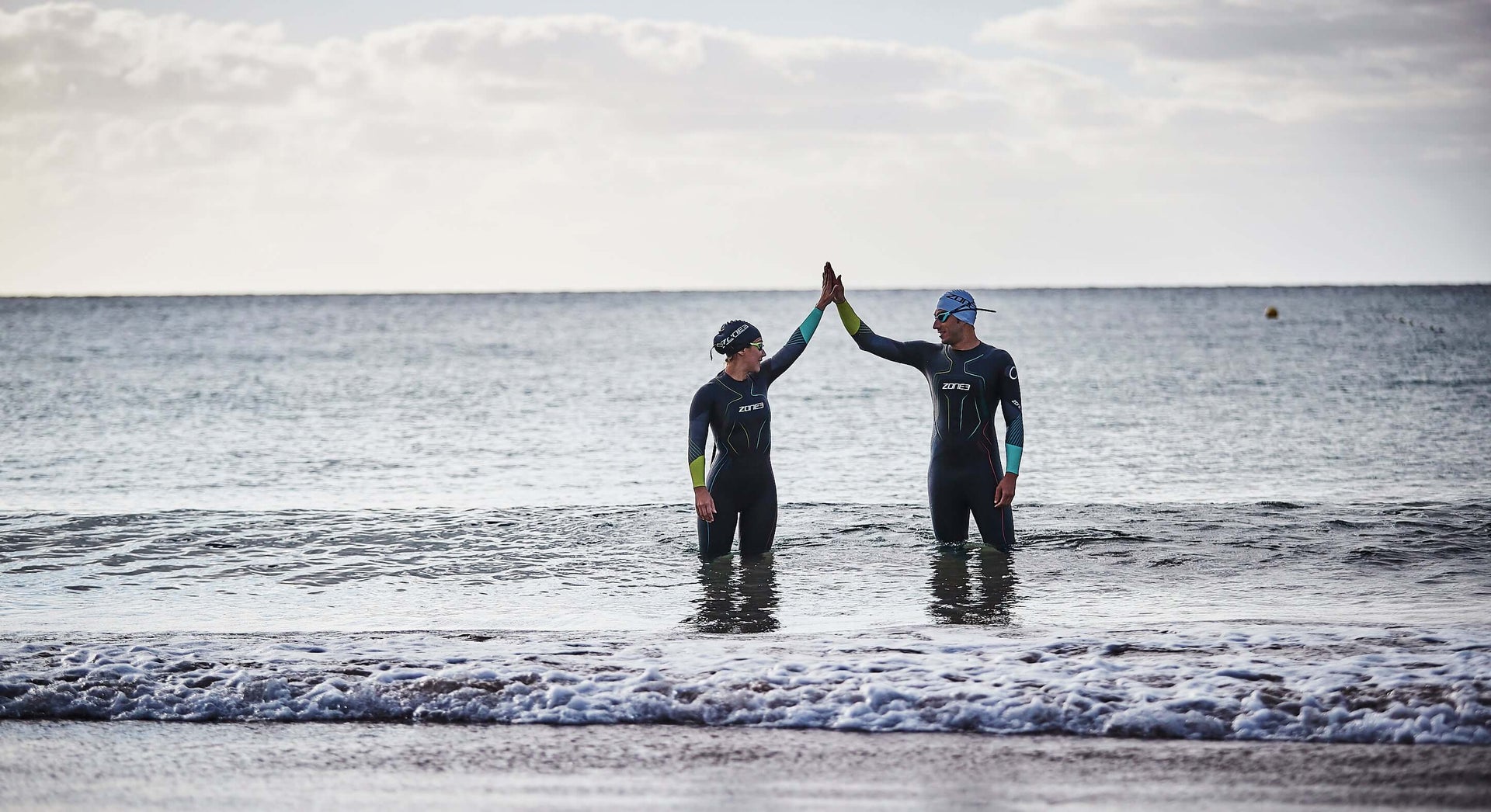 Two triathletes in Zone3 wetsuits high five in shallow ocean water at the beach