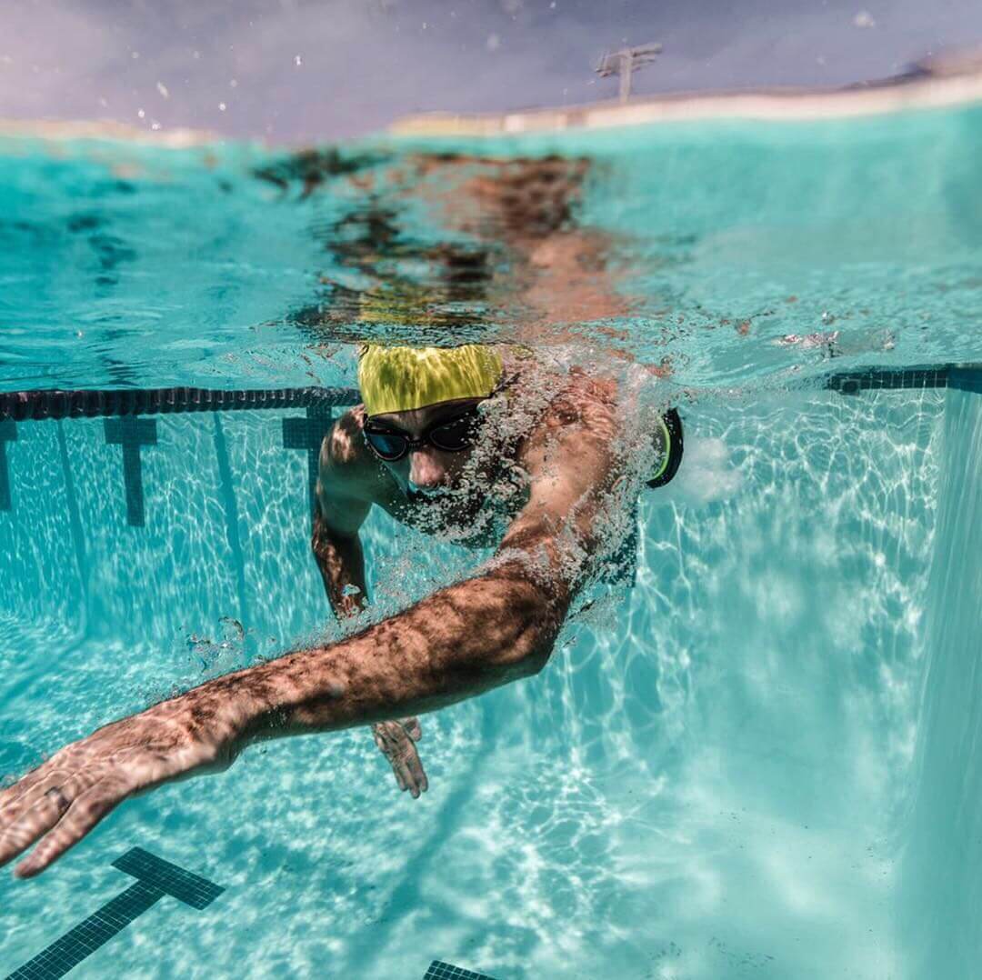 Underwater view of a swimmer in a yellow cap and goggles moving forward beneath the surface of a blue pool.
