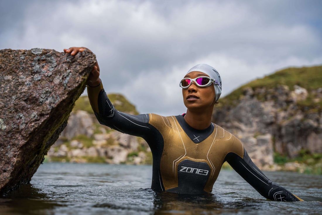 Athlete in a gray neoprene swim cap and mirrored goggles grips a rock on the shore during an open water swim.