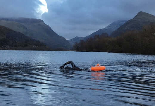 Swimmer in a wetsuit glides across a dark lake with a bright orange buoy and distant mountains.