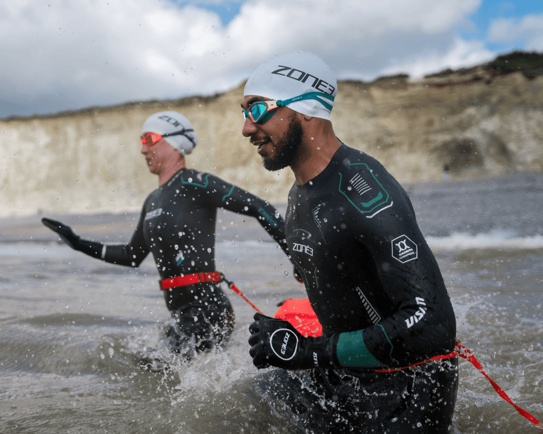 Two swimmers in ZONE3 wetsuits wading out of the sea below white chalk cliffs, one carrying a red tow float.