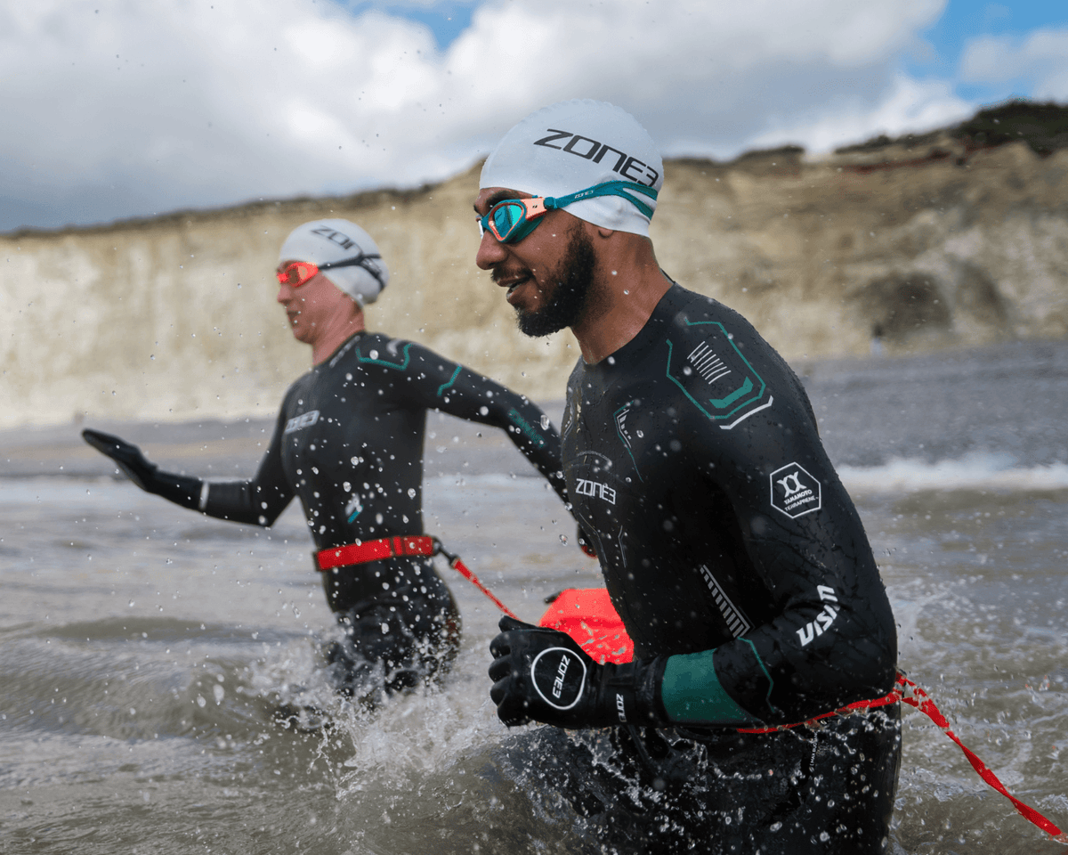 Two swimmers in ZONE3 wetsuits wading out of the sea below white chalk cliffs, one carrying a red tow float.