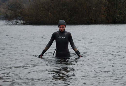 Person in a wetsuit and beanie stands chest-deep in a cold lake ready to swim.