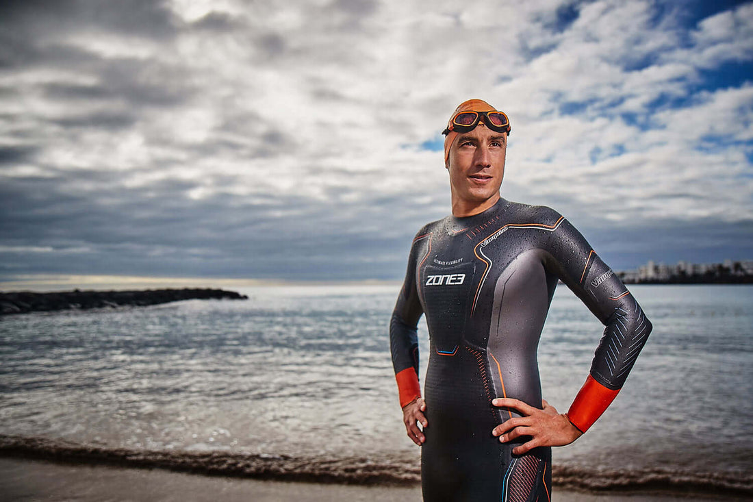 Male triathlete in a Zone3 wetsuit with orange swim cap and goggles on his head stands on a beach by the sea