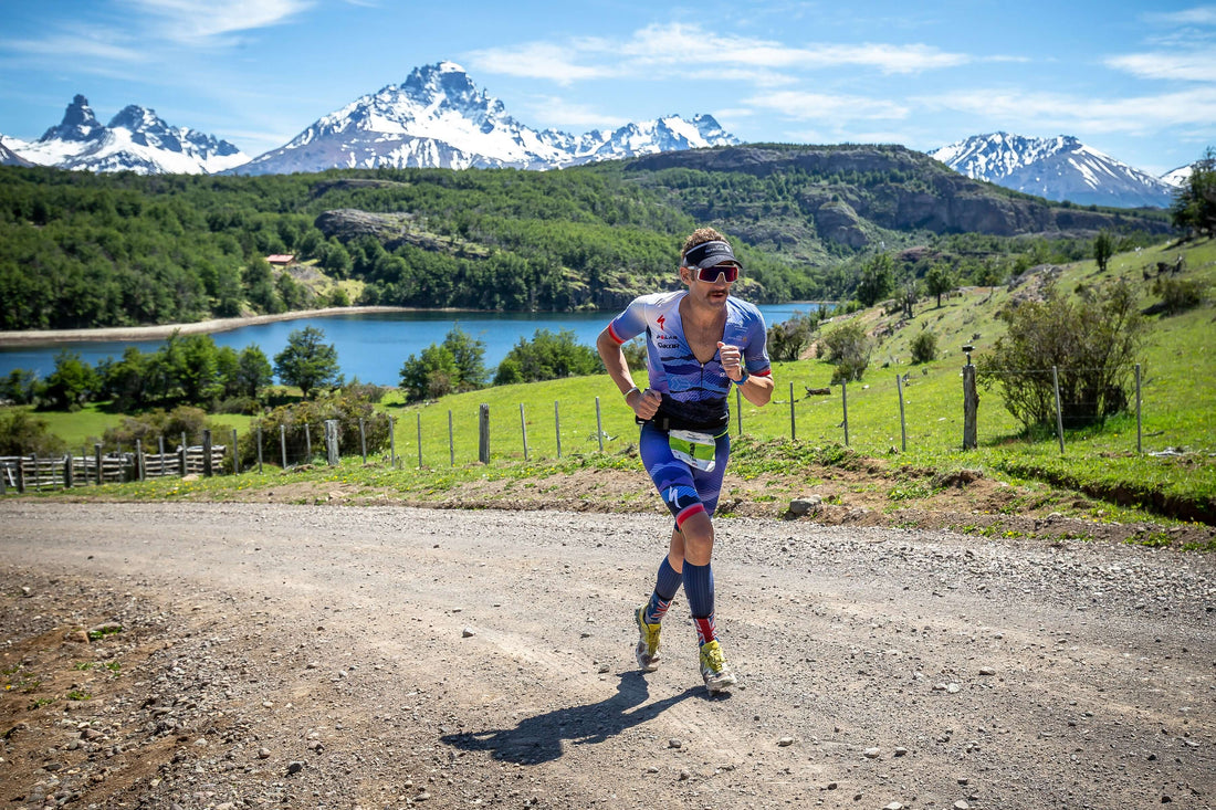 Triathlete in a blue racing suit runs along a gravel road with snow covered mountains and a lake behind.