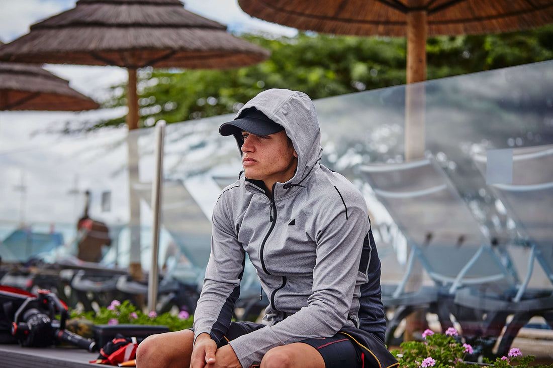 Athlete in a gray hoodie sits outdoors at a race venue with umbrellas and bikes in the background.
