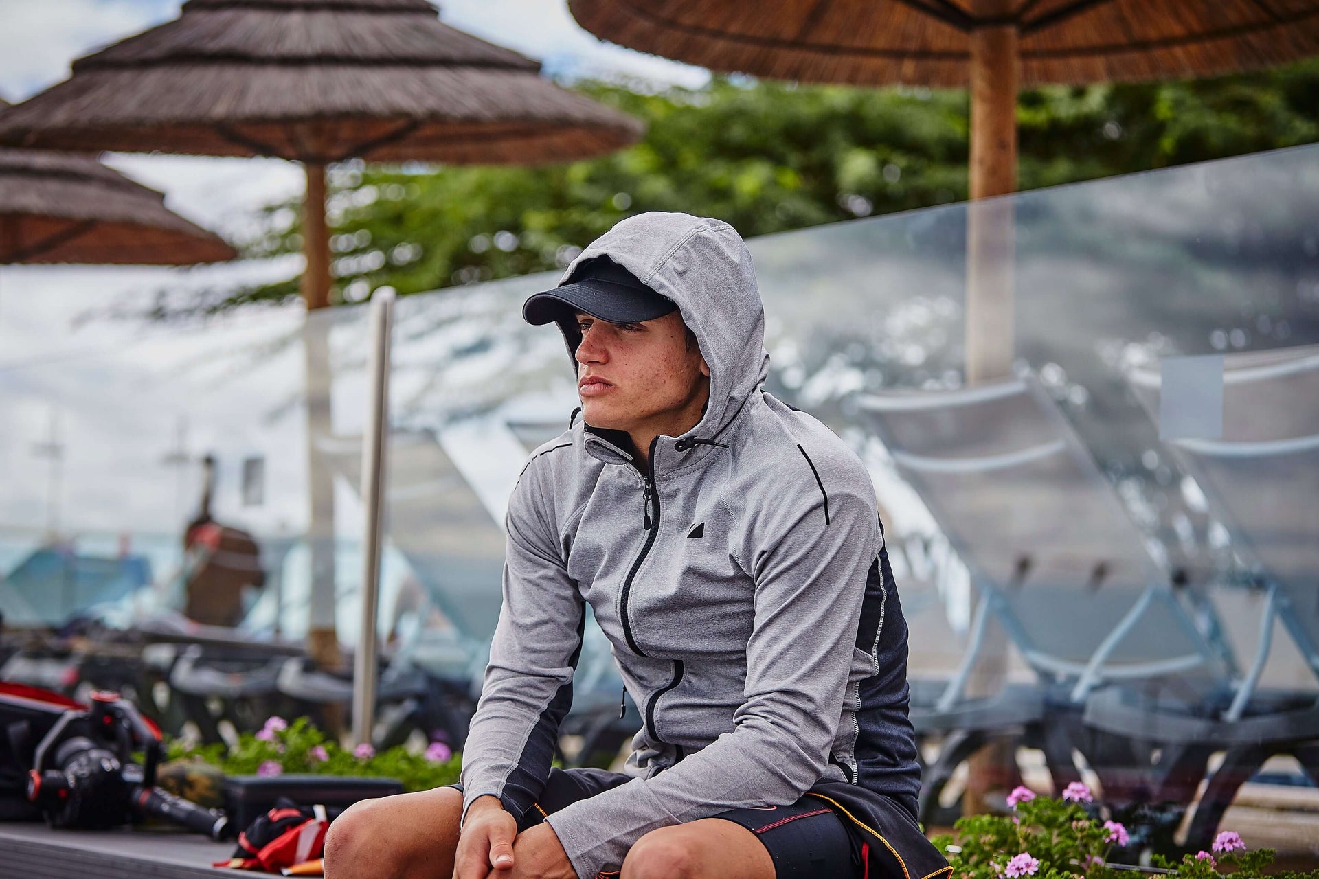 Athlete in a gray hoodie sits outdoors at a race venue with umbrellas and bikes in the background.