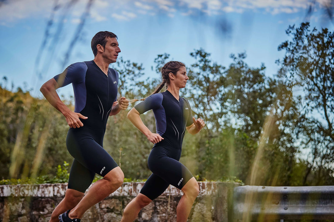 Two runners in matching dark athletic gear sprint along a rural trail with trees and blue sky.
