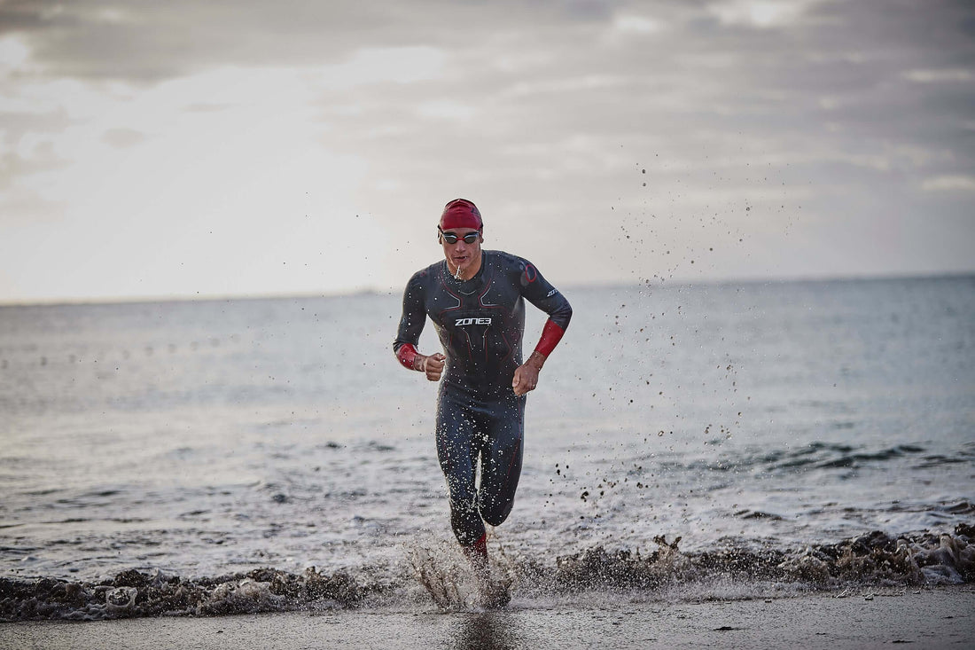 Triathlete runs out of the water wearing a wetsuit a red swim cap and goggles