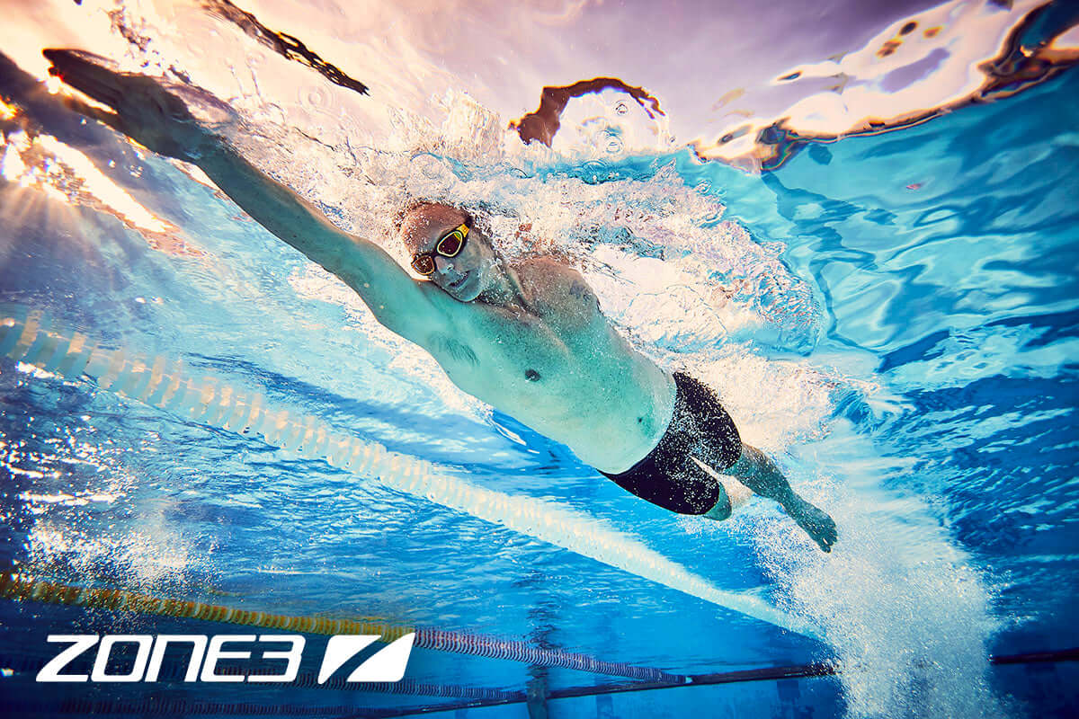 Underwater shot of a male swimmer wearing goggles gliding along a pool lane with Zone3 branding