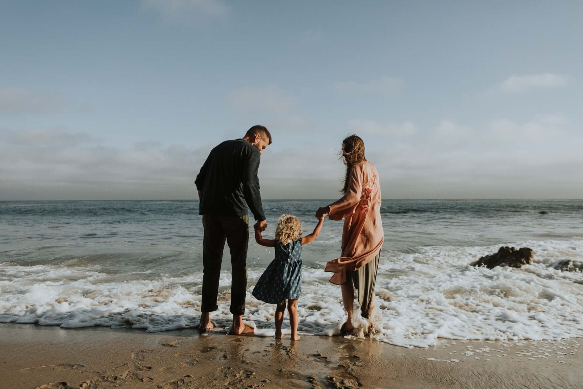 Family of three walking into the sea holding hands on a sandy beach.