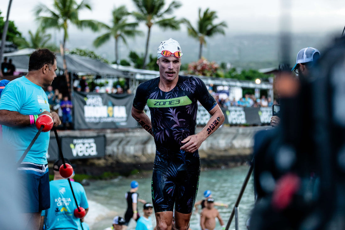 Triathlete in a dark tropical kit with a white cap and red mirrored goggles runs toward the camera at a race setup with volunteers.