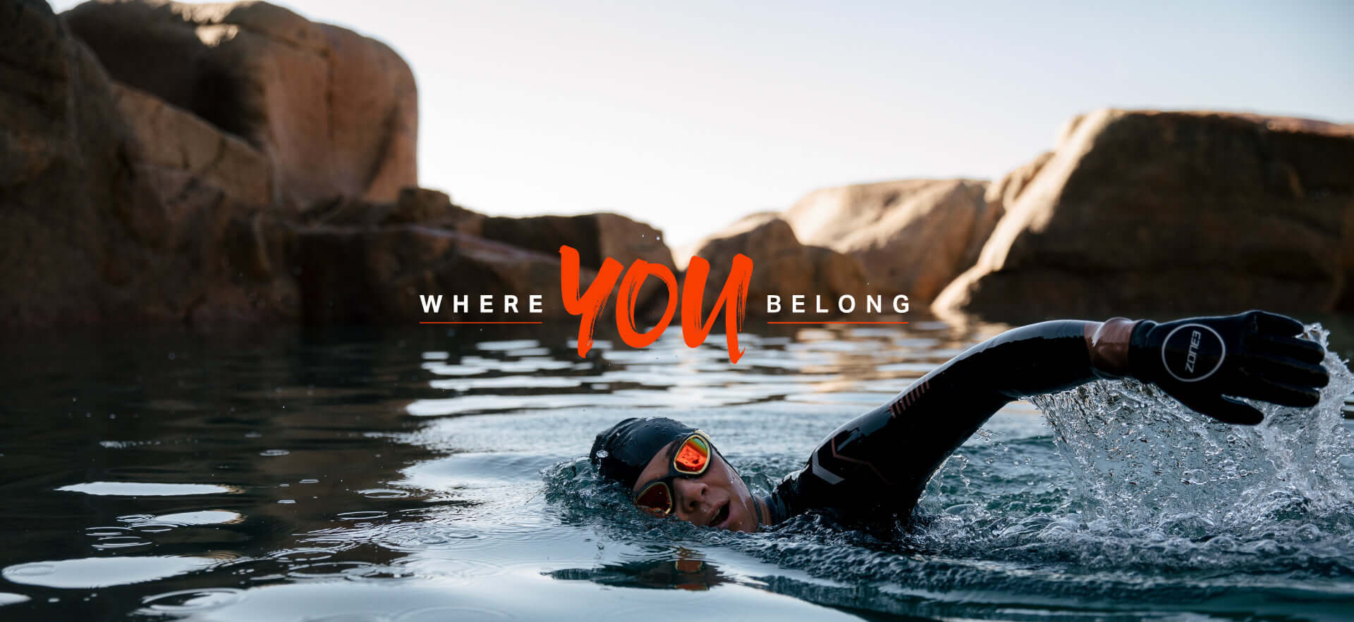 Swimmer in a wetsuit and goggles navigates calm water near rocky cliffs with an orange YOU text overlay