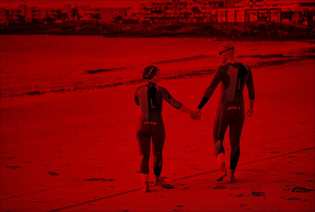 Two athletes in matching triathlon suits walk hand in hand along a beach by the water