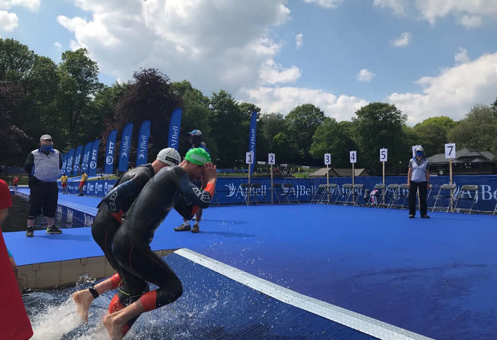 Two wetsuit-clad triathletes dive into the water from a blue transition platform at Leeds WTS with numbered signs in the background