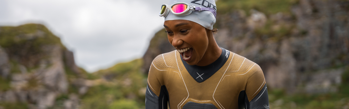 a woman is smiling wearing a gold zone3 wetsuit and swim cap and pink mirrored swim goggles 