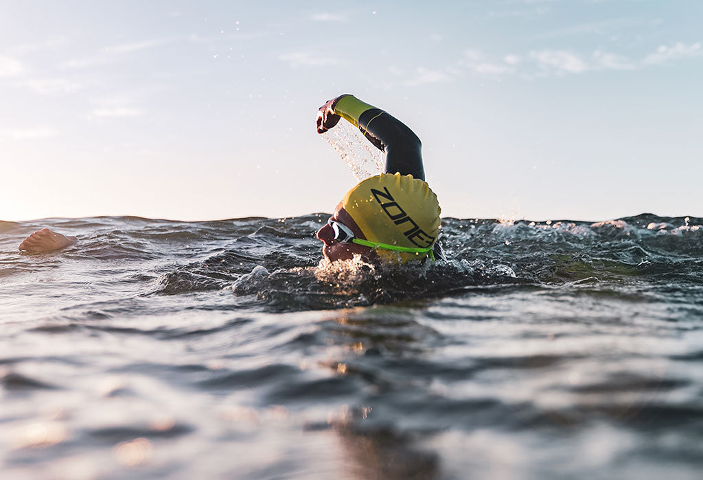 a man is swimming in the sea breathing to one side wearing a zone3 swim cap and goggles