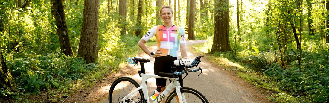 Kaitlin Carew is standing in her trisuit next to her Liv bike on a road surrounded by tall trees on a sunny day