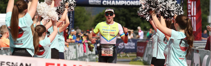 alex lambert is running down the finish line at the Lakesman triathlon, he is wearing a trisuit with sponsors logos on it and sunglasses, people with pompomps are cheering him on