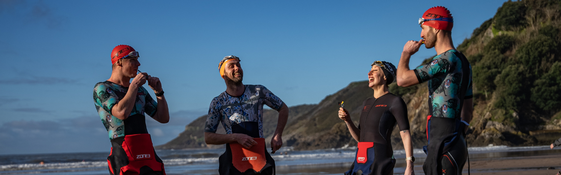 a group of triathletes are stood on the beach in their wetsuits and trisuits laughing and having fun