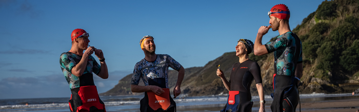 a group of triathletes are stood on the beach in their wetsuits and trisuits laughing and having fun