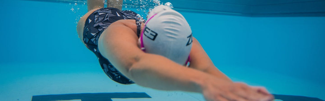 a woman in a zone3 swimming costume, cap and goggles is diving into a swimming pool, she is underwater and the camera angle is also under water