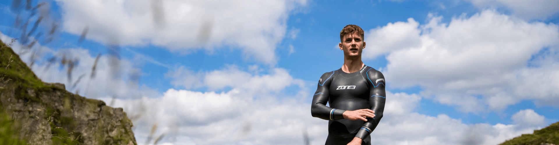 Male athlete in a black and blue Zone3 wetsuit standing outdoors with a blue sky and rocky landscape.