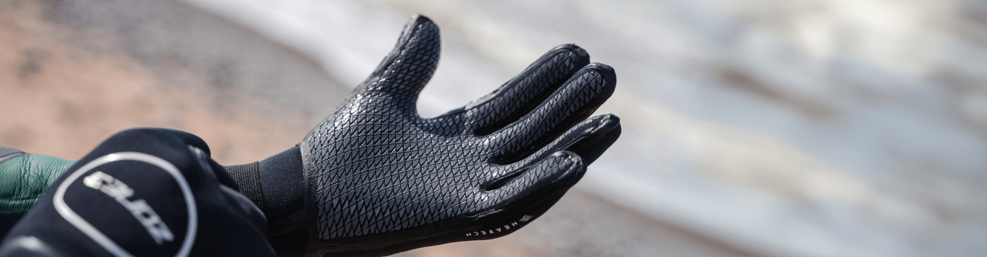 Hand wearing a textured black neoprene swim glove from the Extra Warmth Swim Kit resting on a wetsuit sleeve at the shoreline
