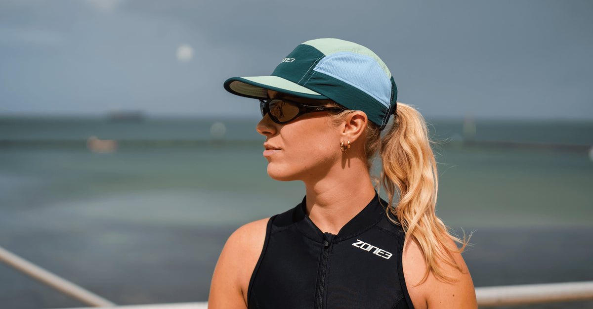 Woman wearing zone3 5 panel running cap and sunglasses with a blurred beach background
