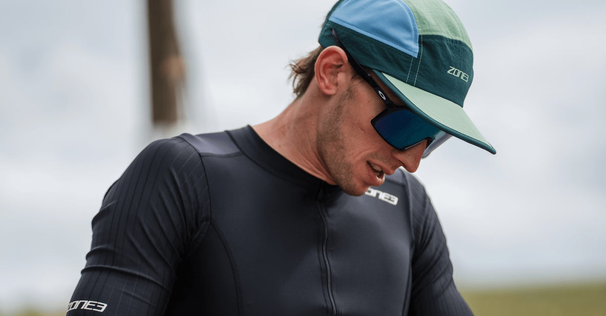 Person wearing black zone3 trisuit with a teal 5 panel running cap and sunglasses, outdoors.