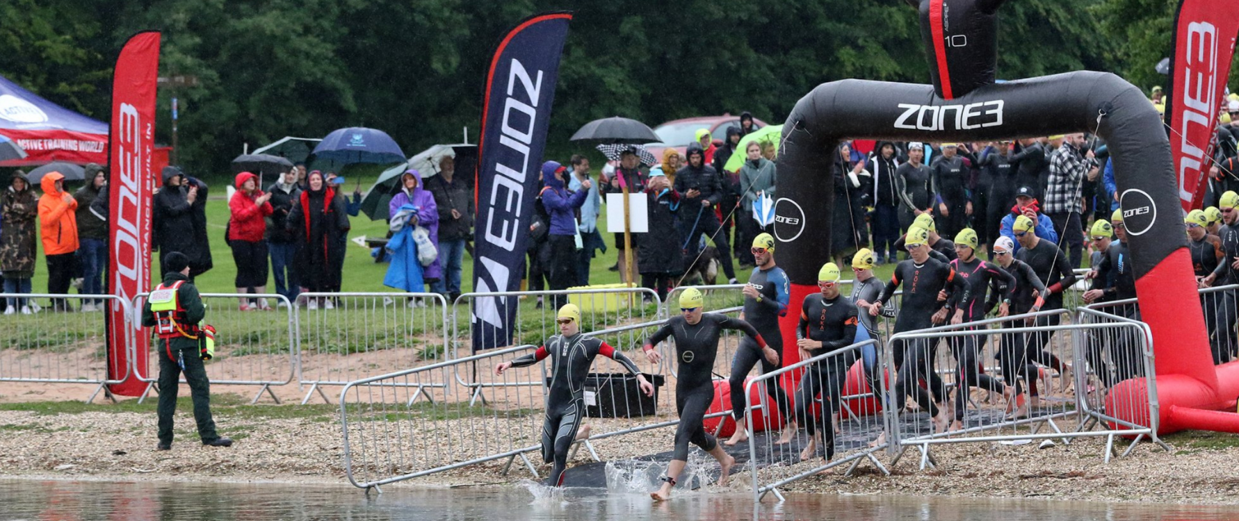 A swim start for a race. Athletes wearing wetsuits running into the water. In the shot is a ZONE3 race gantry, ZONE3 flags and a large crowd of spectators.