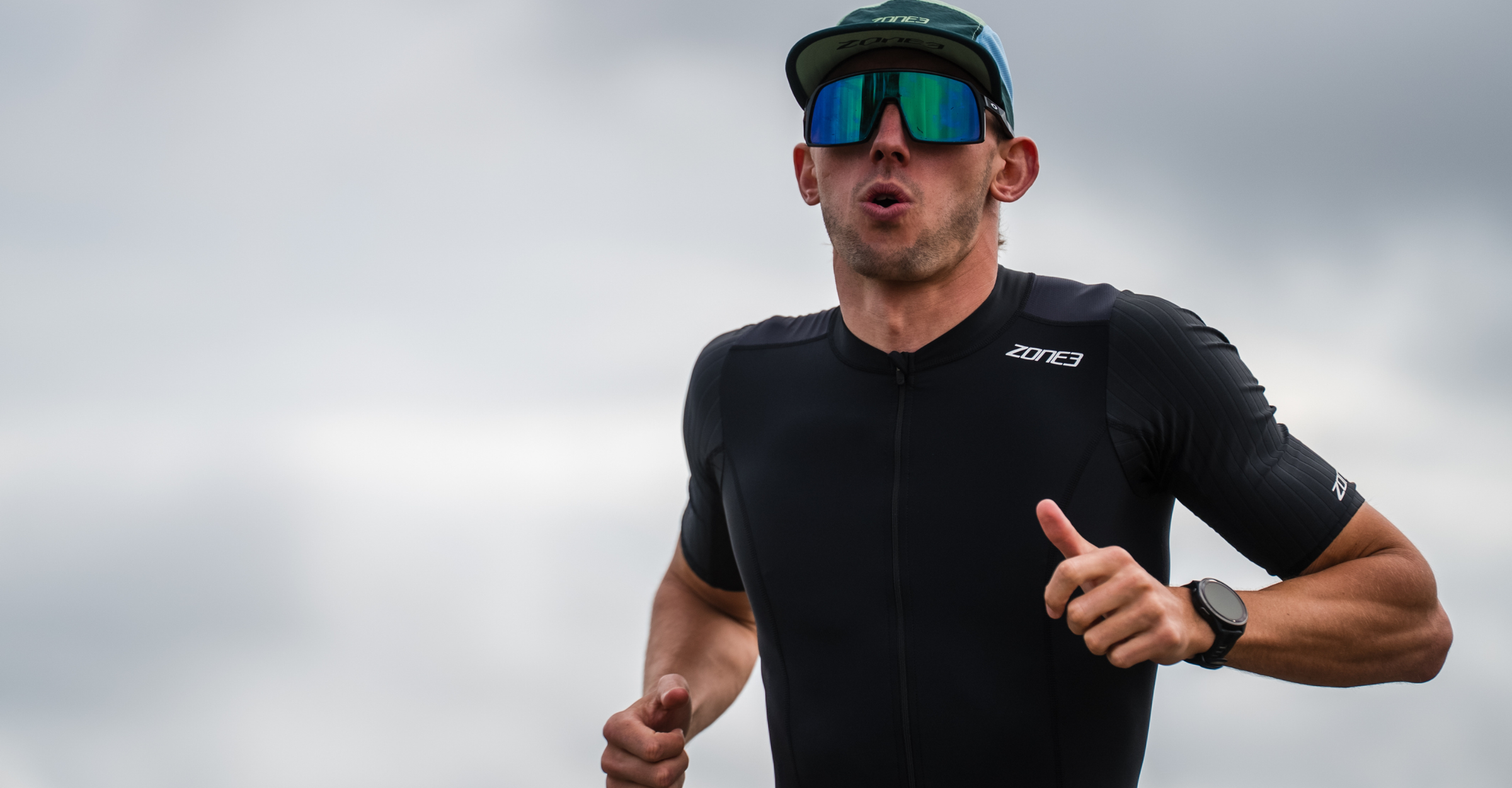 Man running outdoors wearing a black zone3 trisuit and sunglasses, giving a thumbs-up.