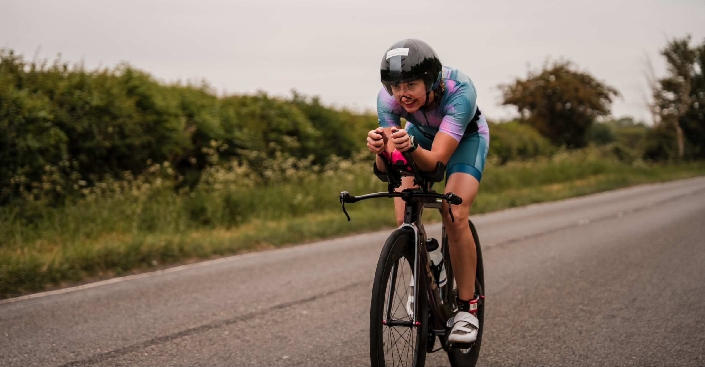 Person in a pink and teal zone3 trisuit riding a bicycle on a road with trees in the background