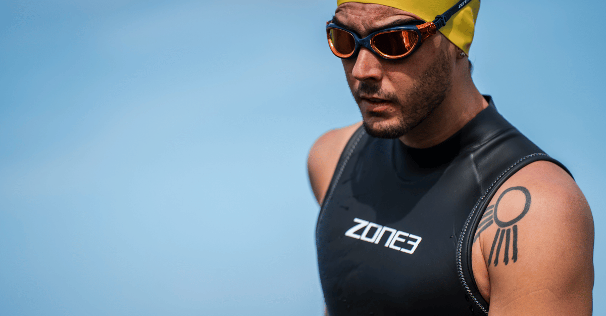 A close-up of a man wearing a black Zone3 Ascend sleeveless wetsuit, yellow swim cap, and orange-tinted goggles against a clear blue sky.