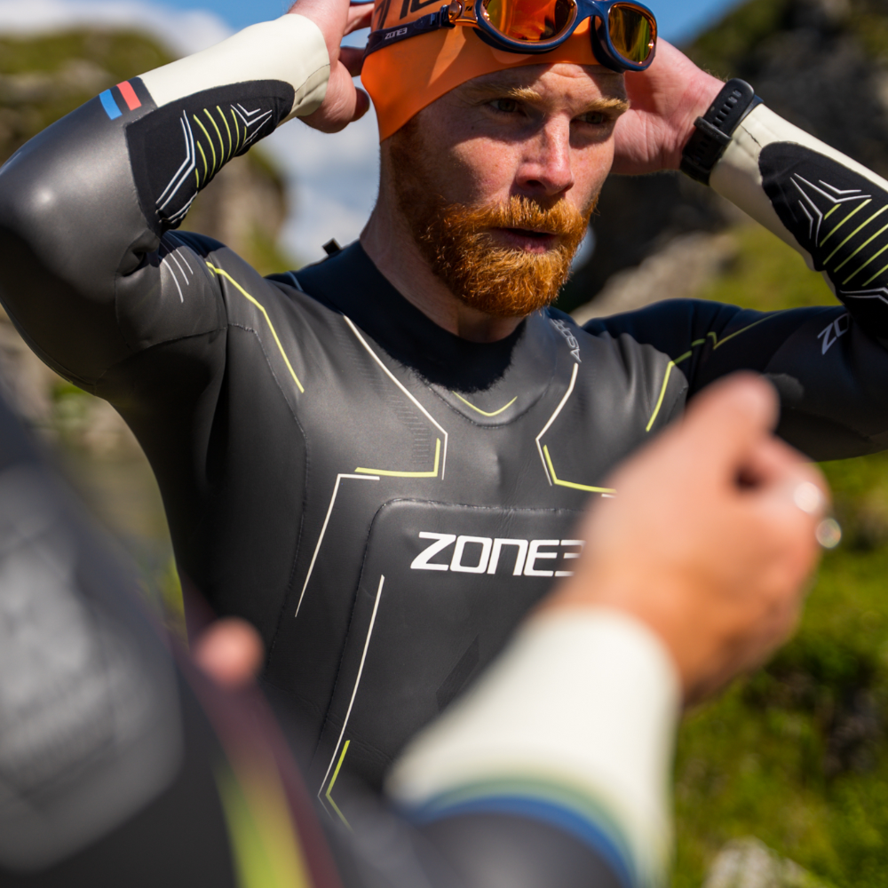 Close up of male wearing a ZONE3 Aspire wetsuit. He is adjusting his orange swim cap and looking into the distance. Background is green and rocky.