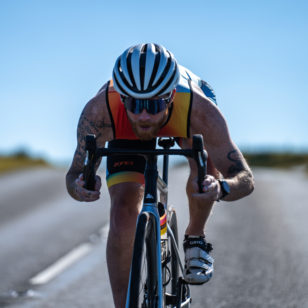 Man wearing a ZONE3 Activate plus trisuit on a bike. He is leaning forward towards the bars of the bike and looking forward.
