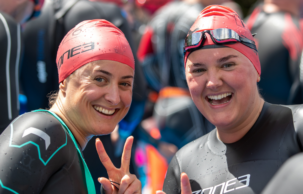 Two athletes in wetsuits and red swim caps smiling and making peace signs.