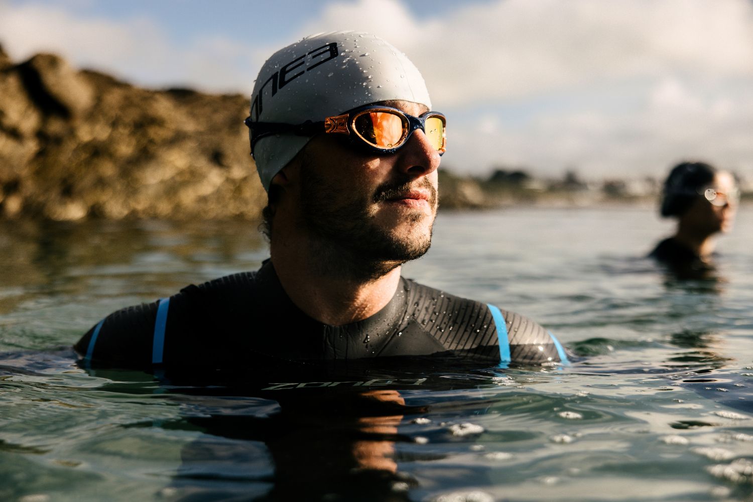 Man wearing a ZONE3 Glide wetsuit, ZONE3 swim cap and ZONE3 Venator-X goggles in a body of water with another person in the background.