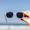 Close-up of a hand holding Waterhaul sunglasses with light-grey frames and dark polarized lenses against a bright blue sky and blurred ocean backdrop. Showcases sustainable, ocean-ready eyewear in a natural coastal setting.