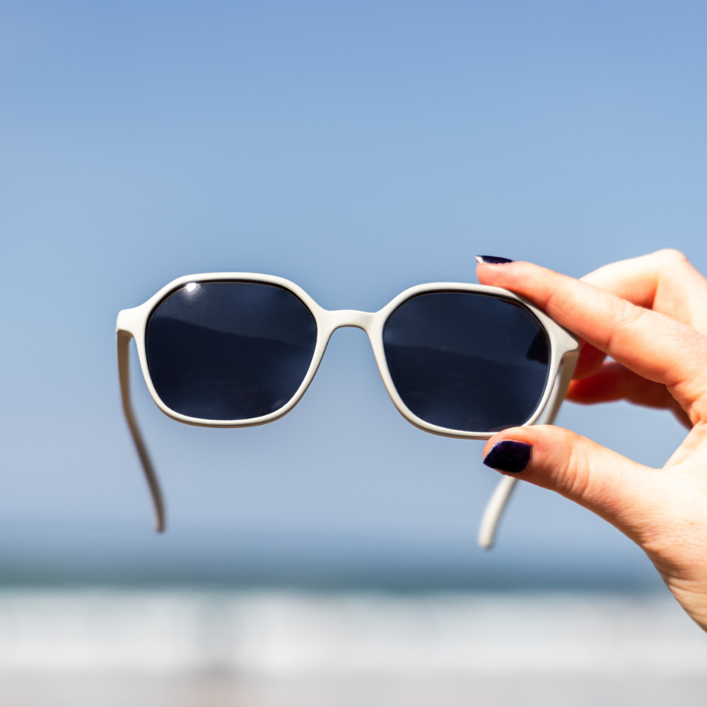 Close-up of a hand holding Waterhaul sunglasses with light-grey frames and dark polarized lenses against a bright blue sky and blurred ocean backdrop. Showcases sustainable, ocean-ready eyewear in a natural coastal setting.