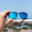 Hand holding Waterhaul sunglasses with matte black frames and blue mirrored polarized lenses, positioned against a bright blue sky and rocky shoreline. Designed for sustainable performance and coastal adventure.