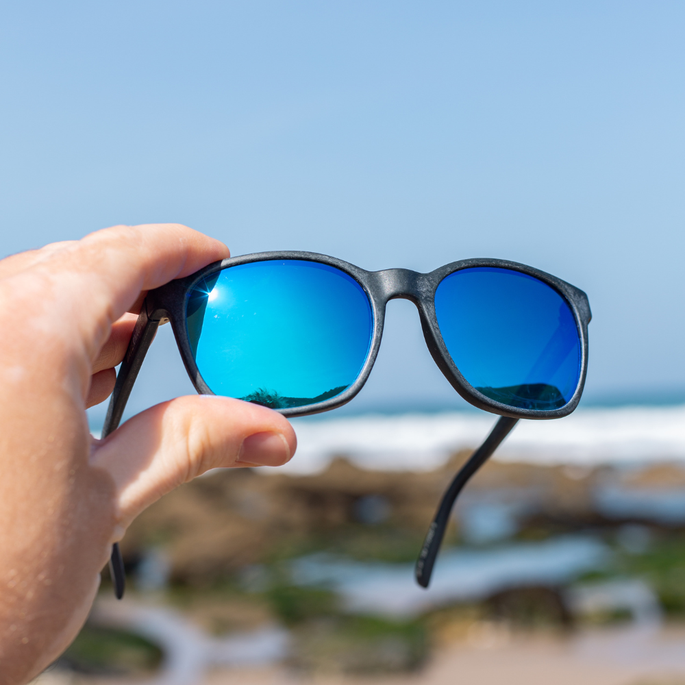 Hand holding Waterhaul sunglasses with matte black frames and blue mirrored polarized lenses, positioned against a bright blue sky and rocky shoreline. Designed for sustainable performance and coastal adventure.