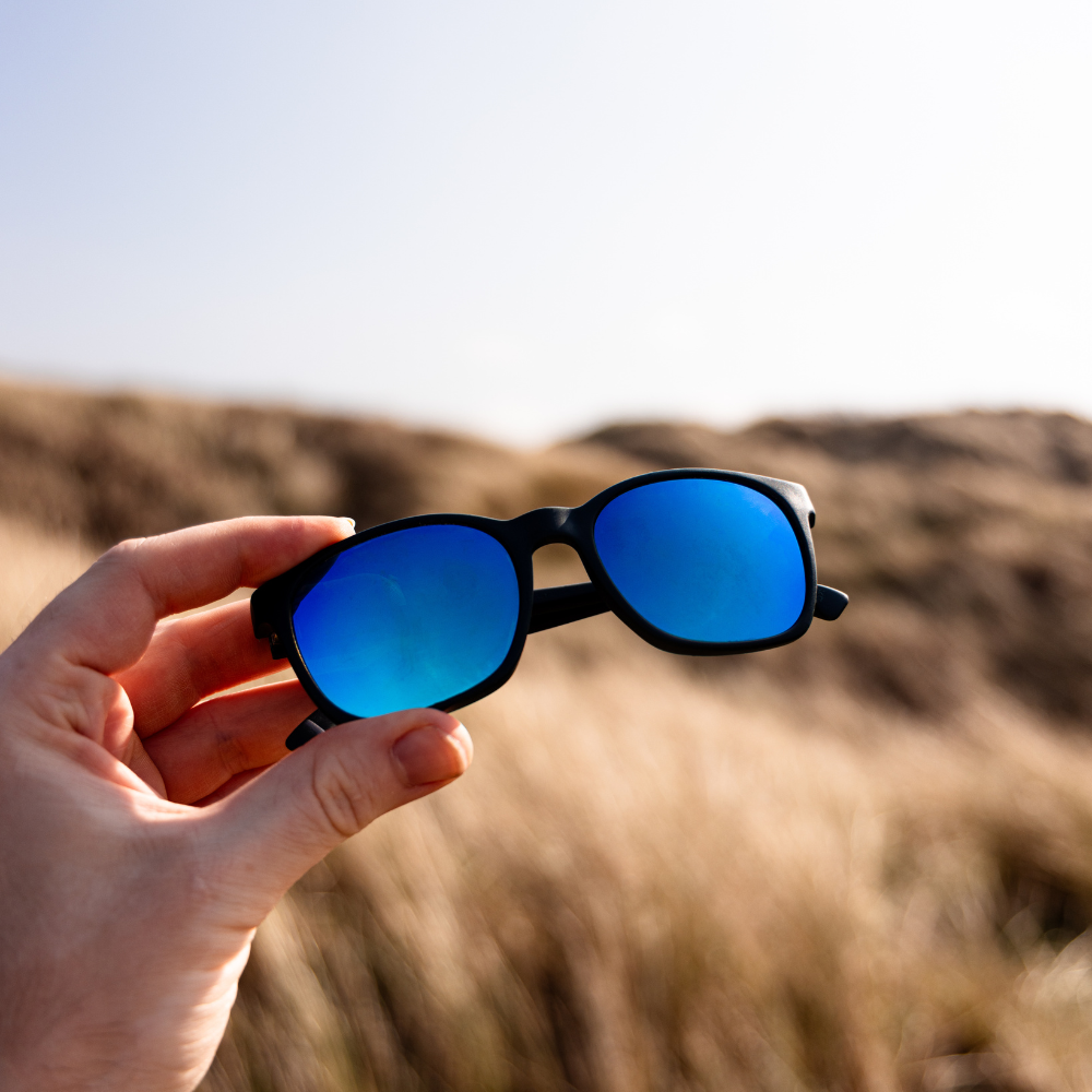 Hand holding a pair of Waterhaul sunglasses with matte black frames and reflective blue polarized lenses, set against a blurred grassy dune landscape under a clear sky. Sustainable eyewear designed for coastal adventure.