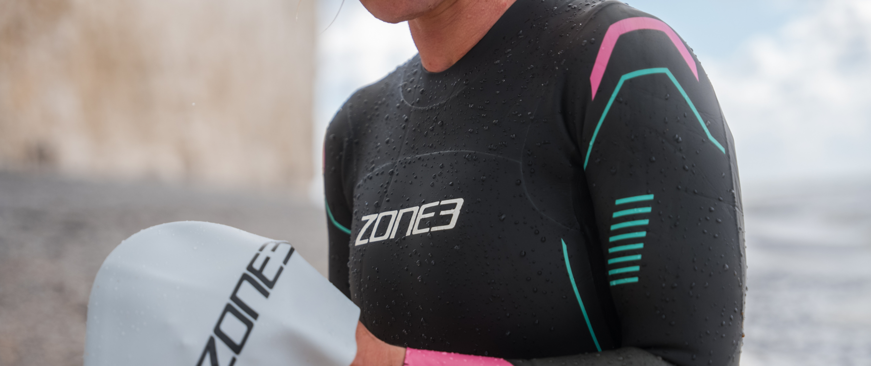 Person wearing a ZONE3 Agile wetsuit with a blurred background. They are holding a ZONE3 Swim cap.