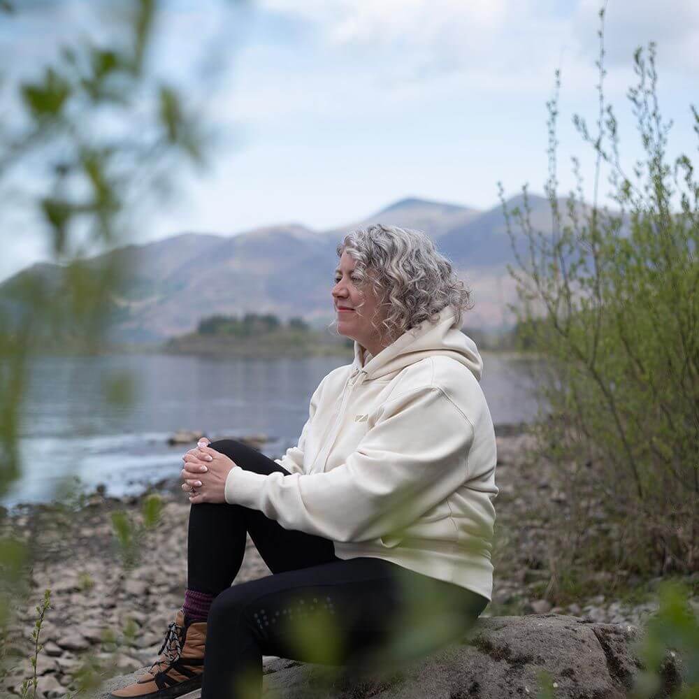 A women wearing the Zone3 Haweswater Hoodie in white sitting on a rock by a lake.