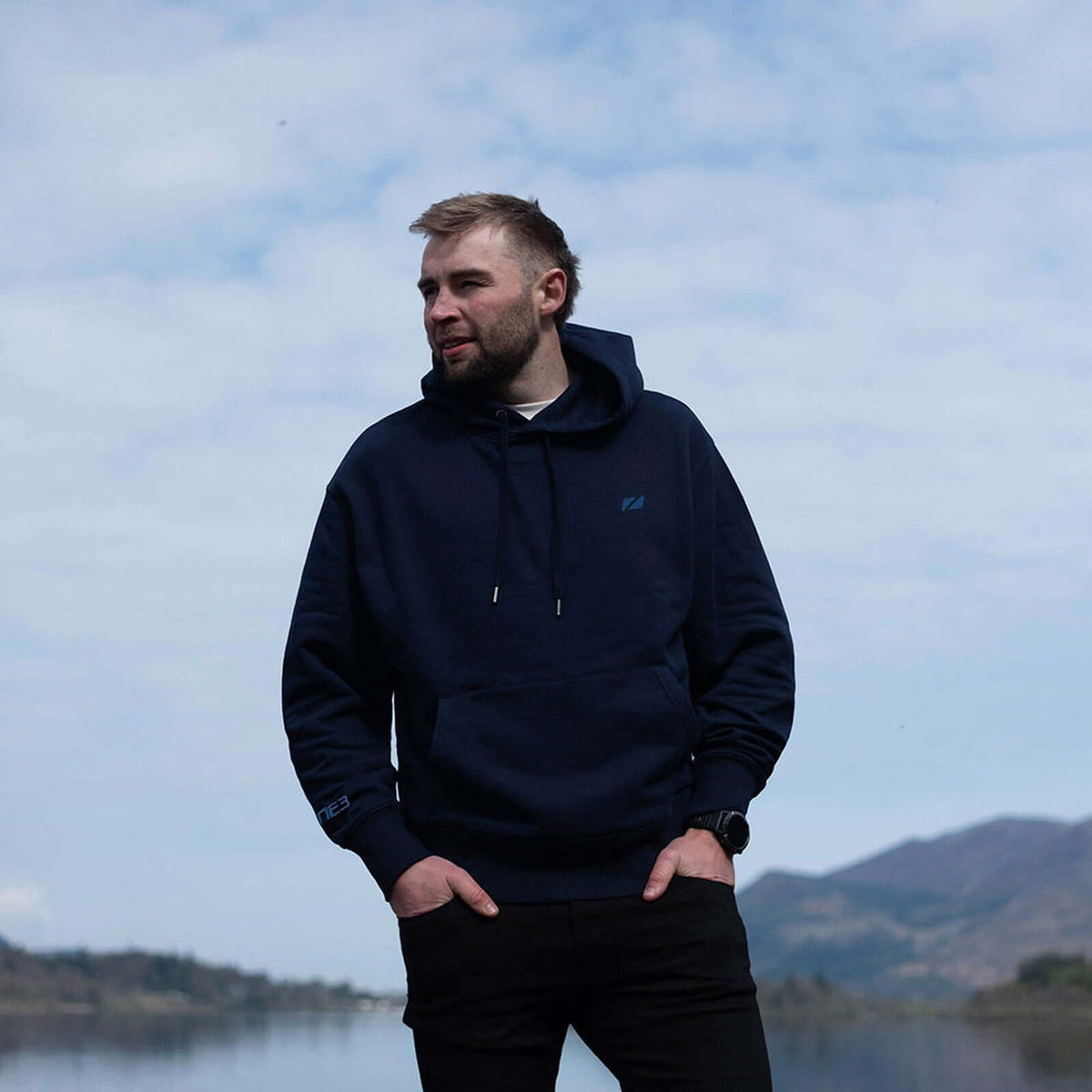A man standing by a lake wearing a Zone3 Haweswater Hoodie in navy. 
