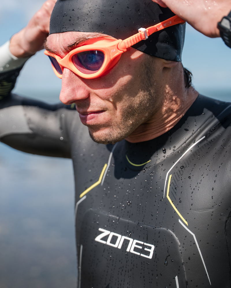 A close-up of a man wearing a black wetsuit with yellow accents, a black swim cap, and orange swim goggles, with water droplets on his skin against an outdoor water background.