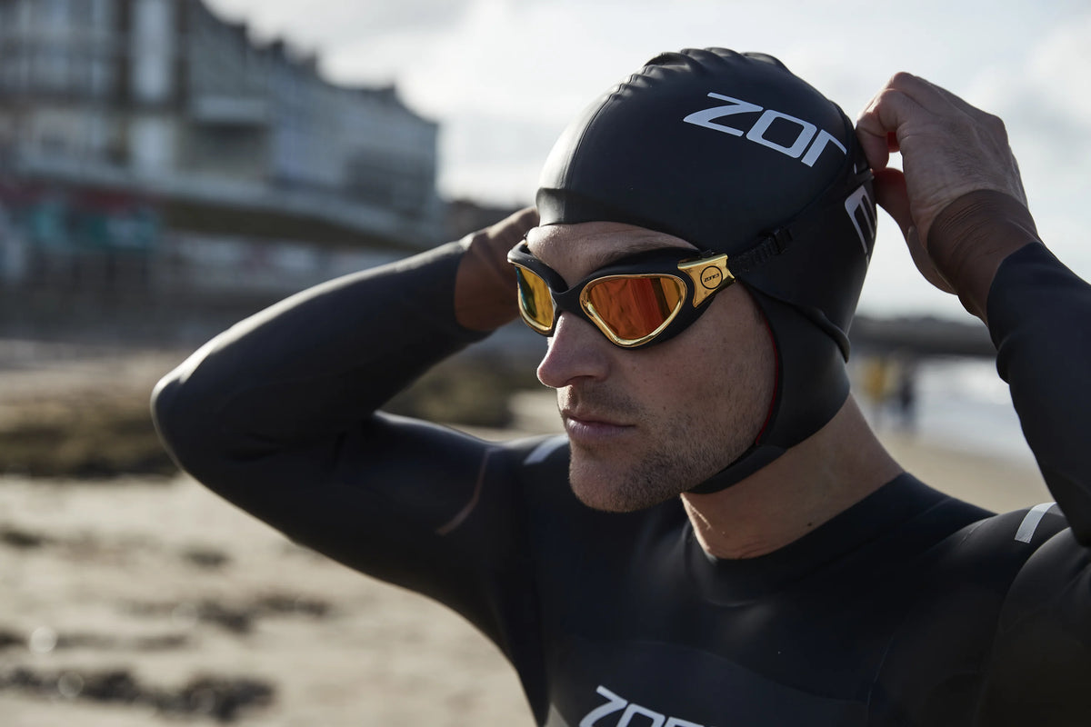 Athlete wearing the Zone3 Heat tech swim cap at the beach preparing for swim as he places a Pair of Zone3 goggles. 