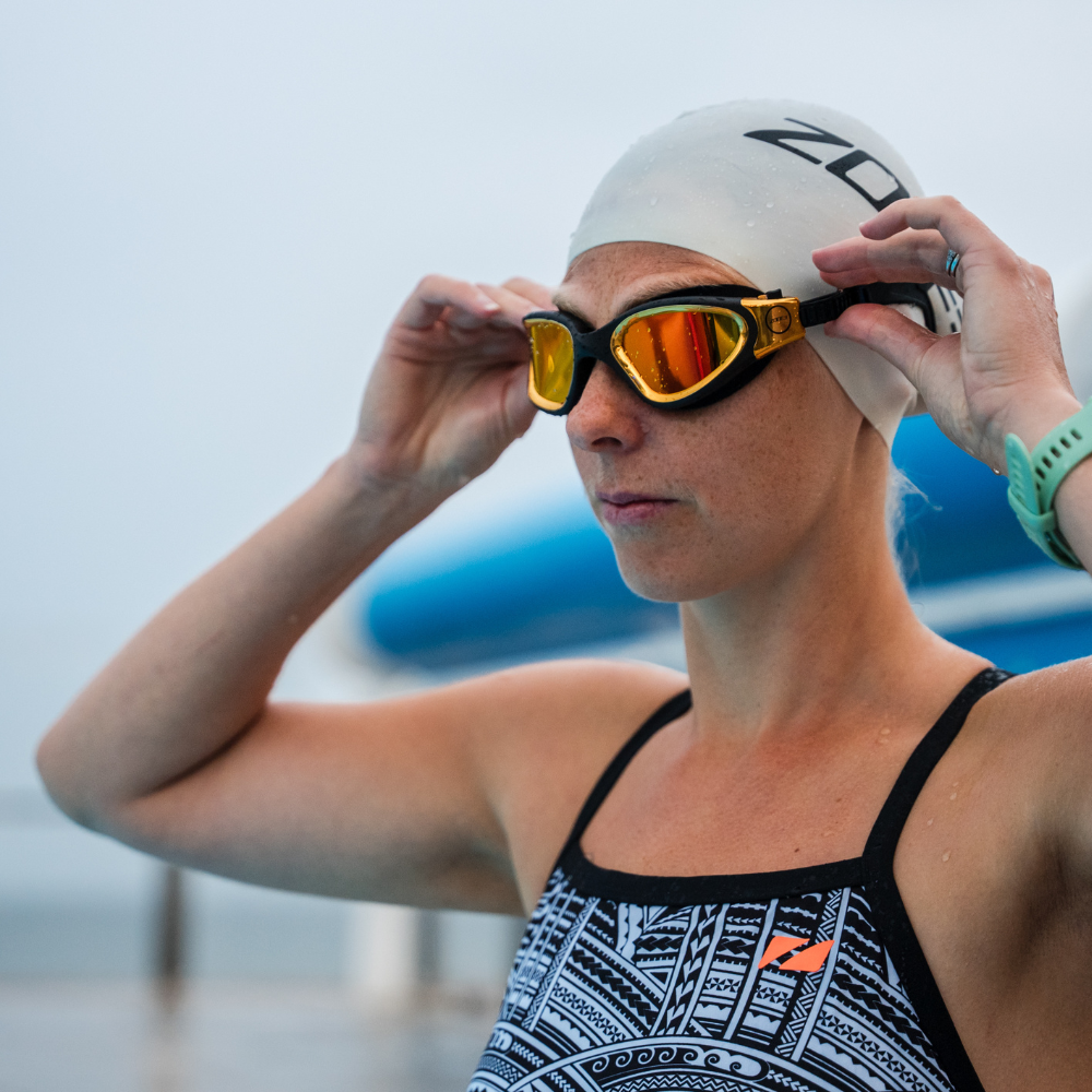 Swimmer wearing a white swim cap and gold mirrored goggles, adjusting the strap. She is wearing the Kona Speed patterned swimsuit with thin black straps.