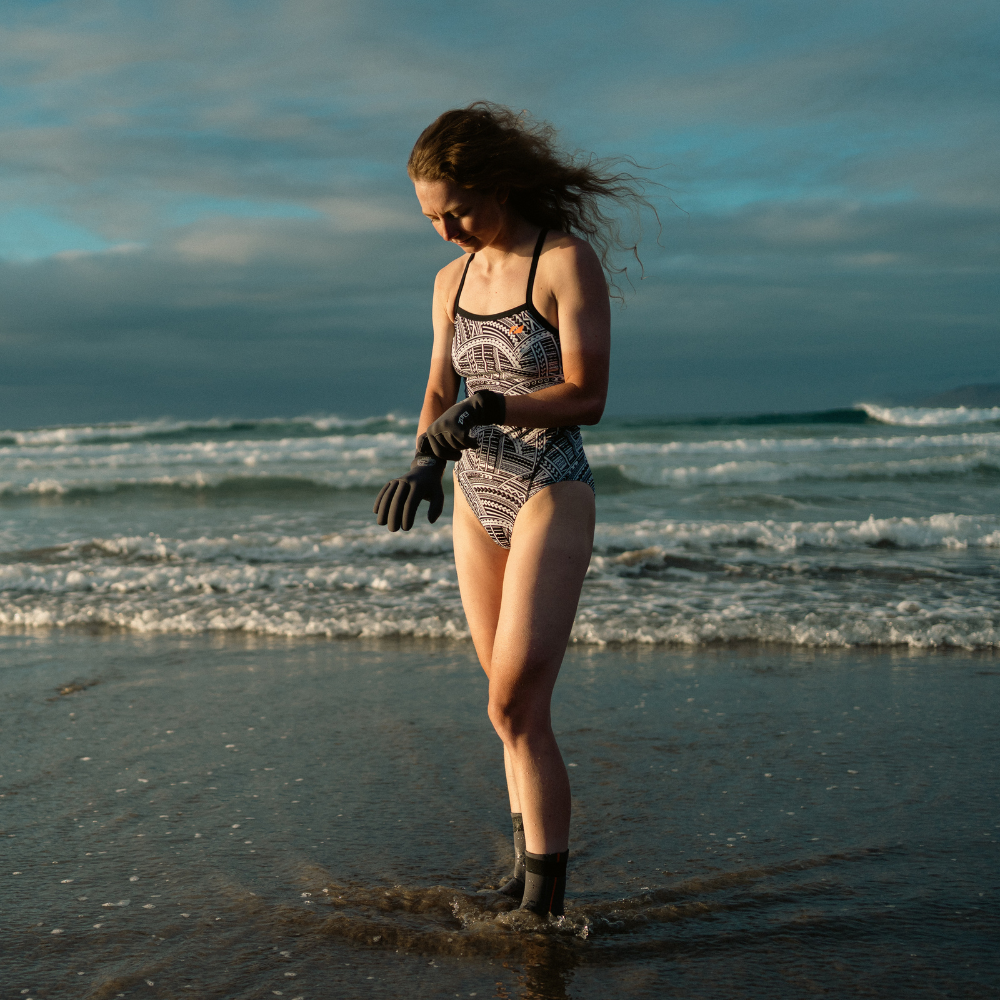 Female swimmer standing at the shoreline wearing the ZONE3 Kona Speed print swimsuit, neoprene swim gloves, and boots. 